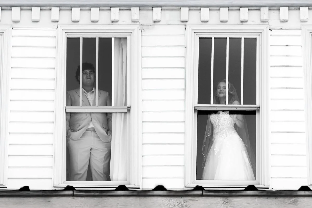 A black and white photo of a man and a woman standing behind separate window screens of a house. The man is dressed in a suit and the woman is wearing a wedding dress, smiling, suggesting a wedding or special occasion.