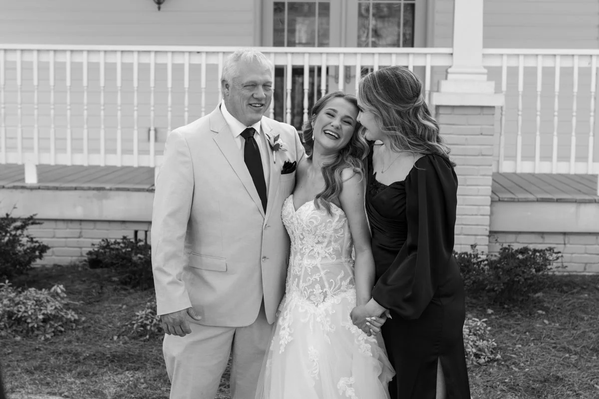 A bride in a wedding dress stands between an older man in a light suit and a bride in a black dress, all smiling and holding hands, outside in front of a house.