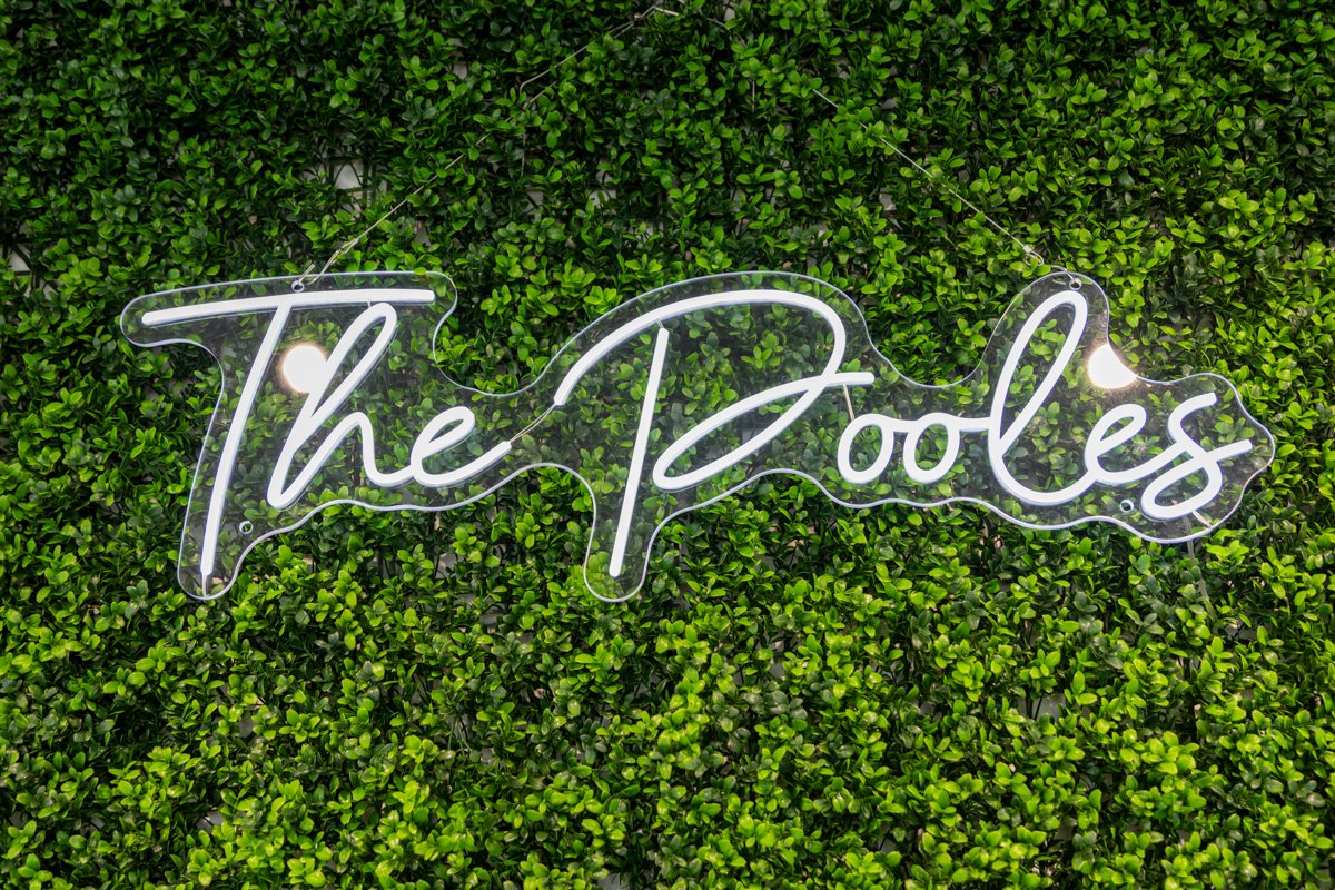 Neon sign reading 'The Pooles' mounted on a green leafy wall.