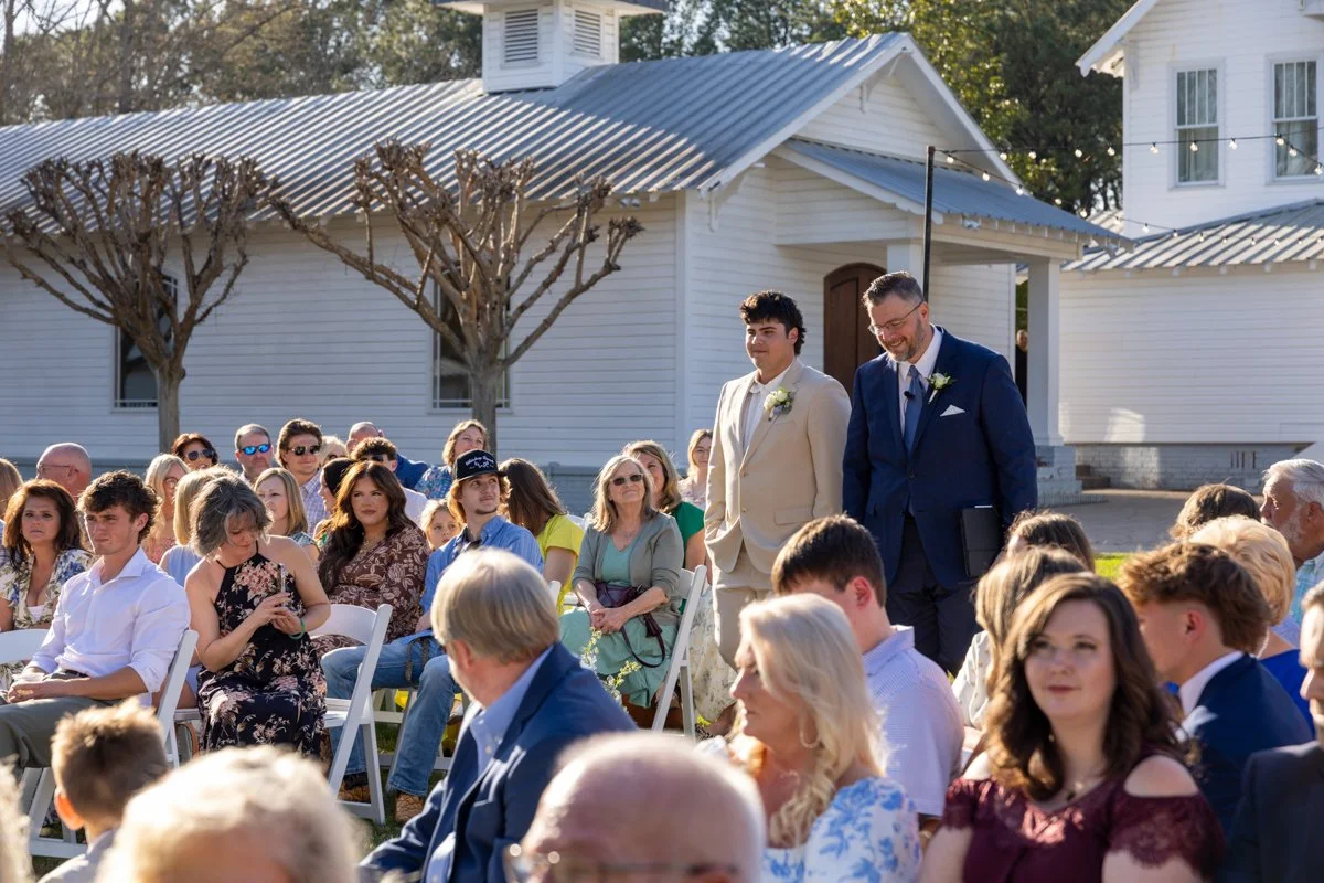 Outdoor wedding ceremony with two men walking down the aisle, one in a beige suit and the other in a dark blue suit, surrounded by seated guests in front of white buildings and leafless trees.