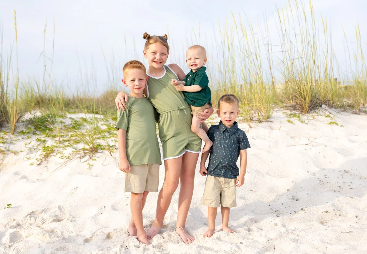 A mother and four children standing on a sandy beach with tall grasses in the background. The mother is holding a toddler while the other three children stand beside her, all smiling and wearing casual summer clothes.