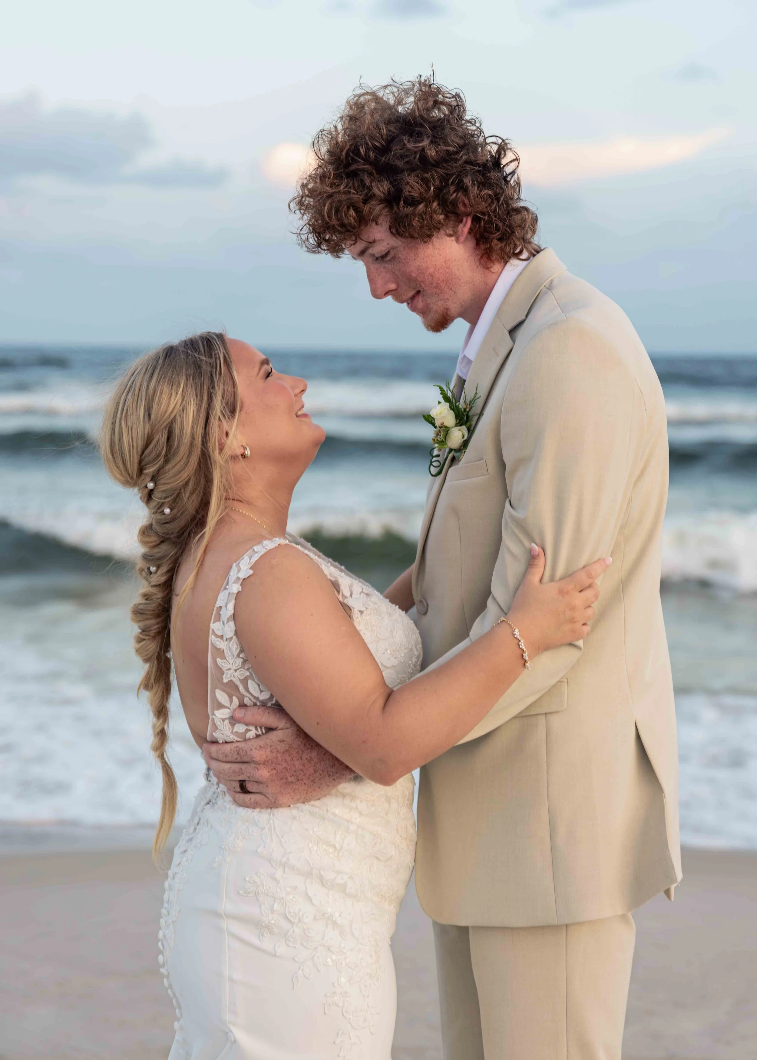 A bride and groom embrace on the beach, the bride wearing a white lace wedding dress and the groom in a light beige suit with a boutonniere. They look into each other's eyes with the ocean and sky in the background.