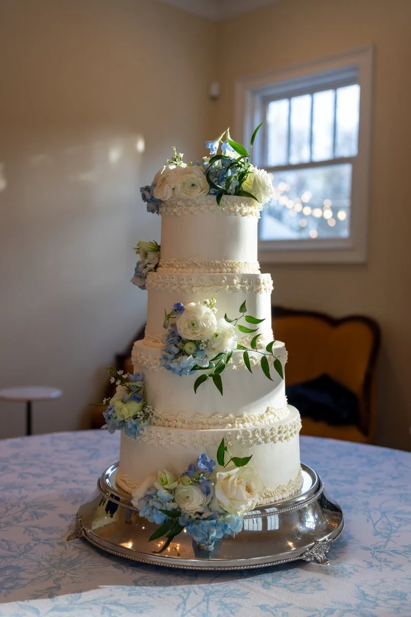 A four-tier white wedding cake decorated with white and blue flowers and green leaves, placed on a silver cake stand on a table with a light blue tablecloth in a room with a window and a yellow armchair in the background.