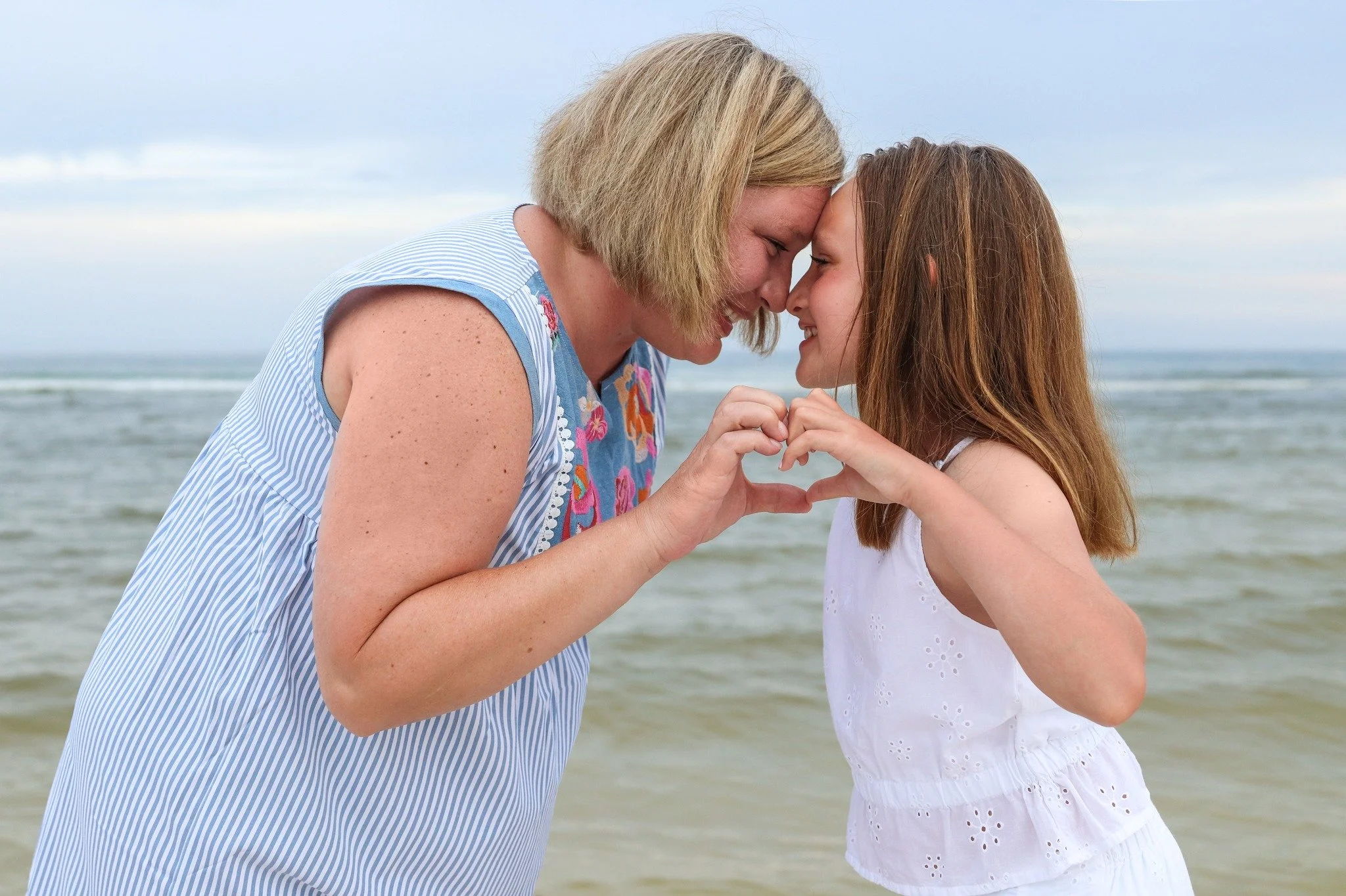 A woman and a girl are facing each other at the beach, touching foreheads and holding hands with fingers interlocked, forming a heart shape. The ocean and cloudy sky are in the background.
