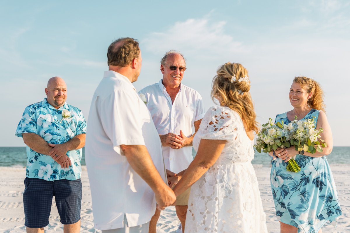 A beach wedding with a couple holding hands and exchanging vows, surrounded by their wedding party and the ocean in the background.