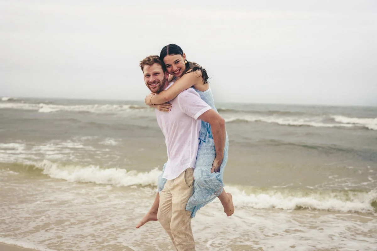 A happy couple at the beach, with the woman riding on the man's back, both smiling.