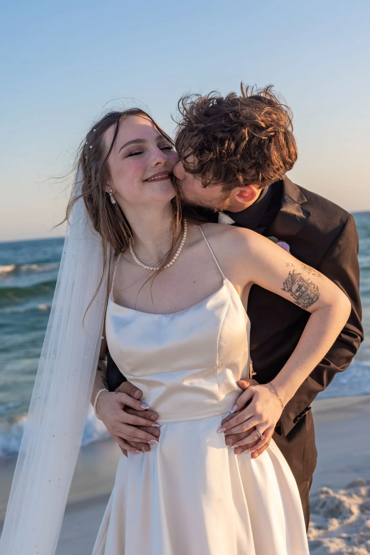 A newlywed couple sharing a kiss on the beach during sunset, dressed in wedding attire, with ocean waves in the background.
