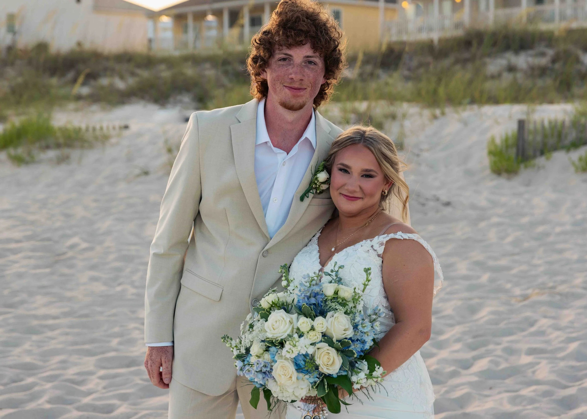 A couple dressed in wedding attire standing on a sandy beach with beach houses in the background, smiling and holding a bouquet of white and blue flowers.