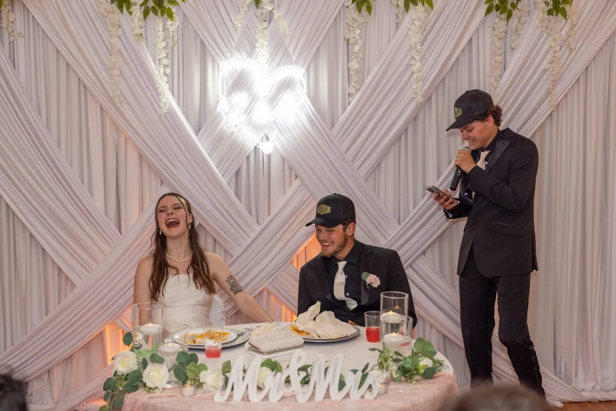 A bride and groom laughing and smiling while sitting at a wedding reception table, with a person giving a speech or toast standing nearby. The background features draped white fabric and soft lighting.