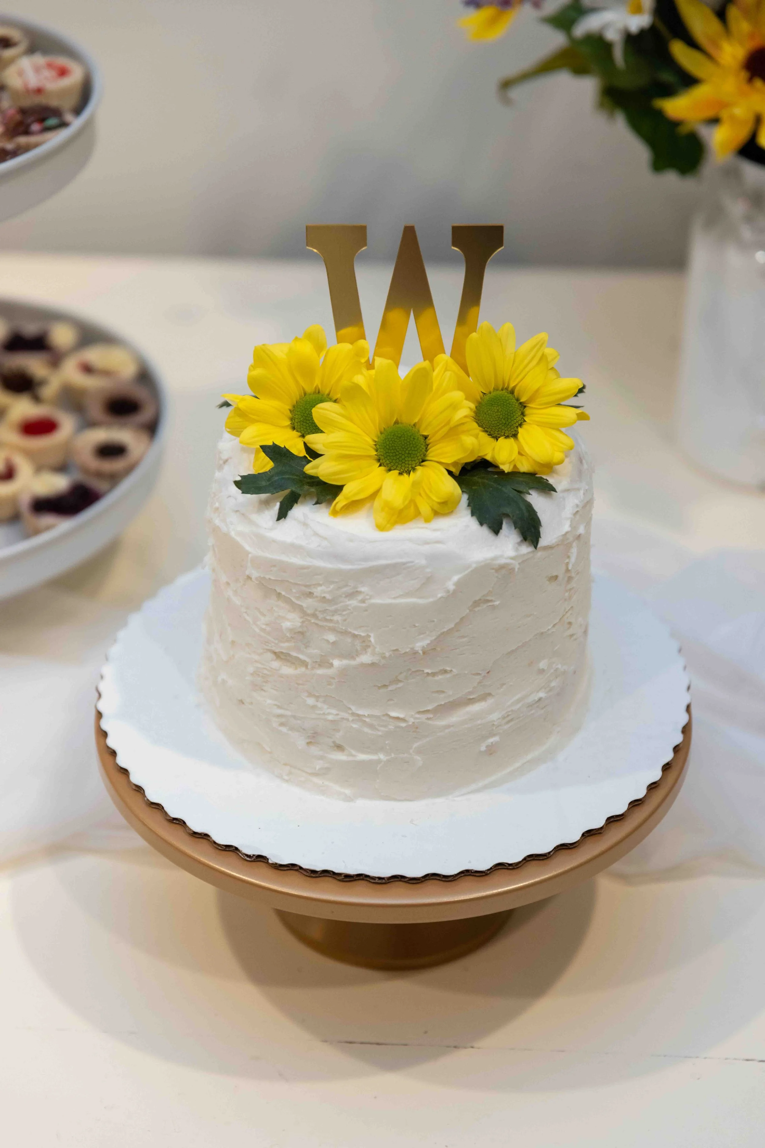A cake decorated with yellow sunflowers and a gold letter W topper, placed on a white stand.