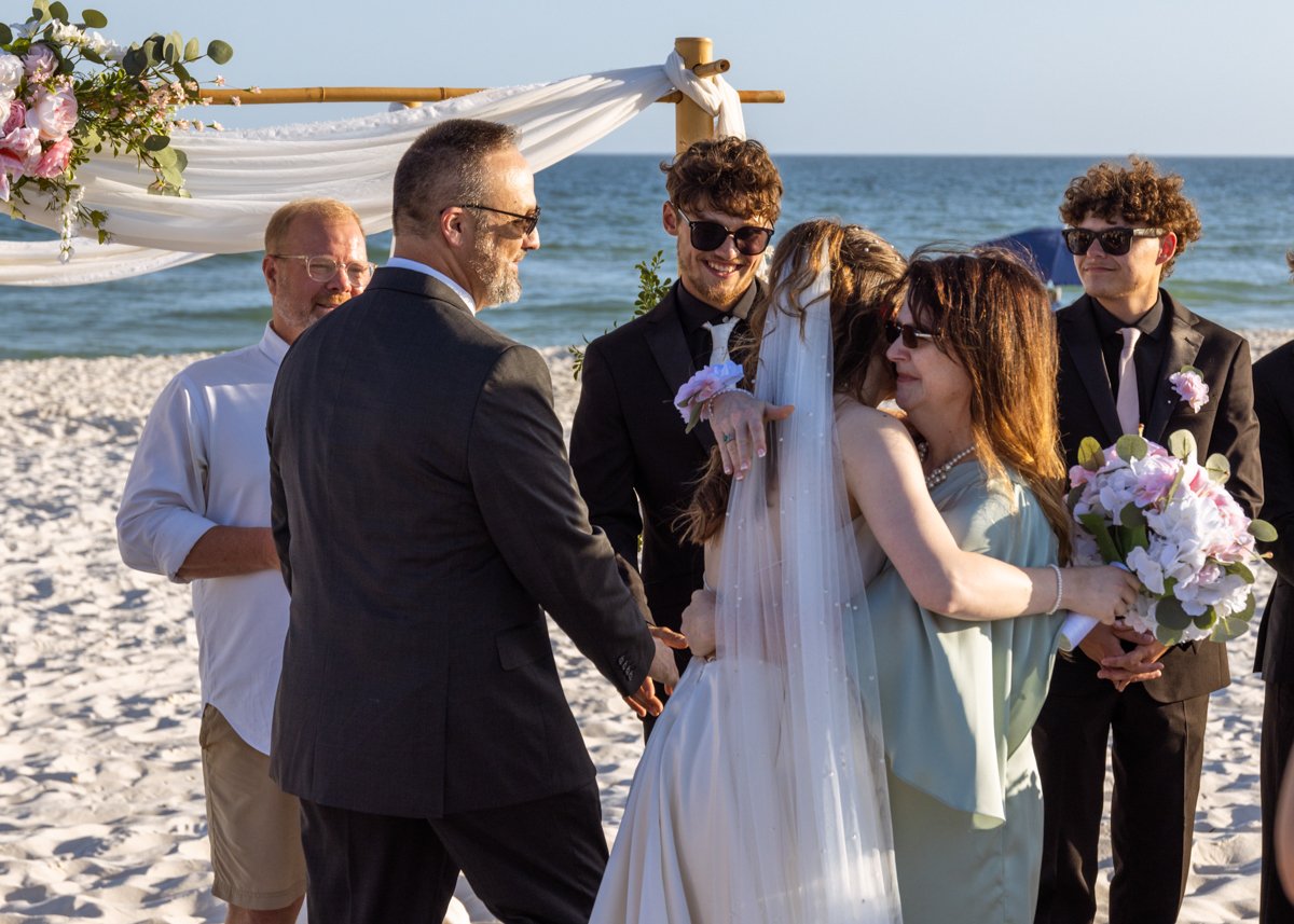 A beach wedding ceremony with a bride and groom exchanging vows, surrounded by friends and family, near the ocean with clear skies.