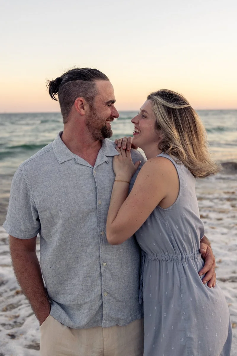 A man and woman smiling at each other on the beach during sunset, with ocean waves in the background.