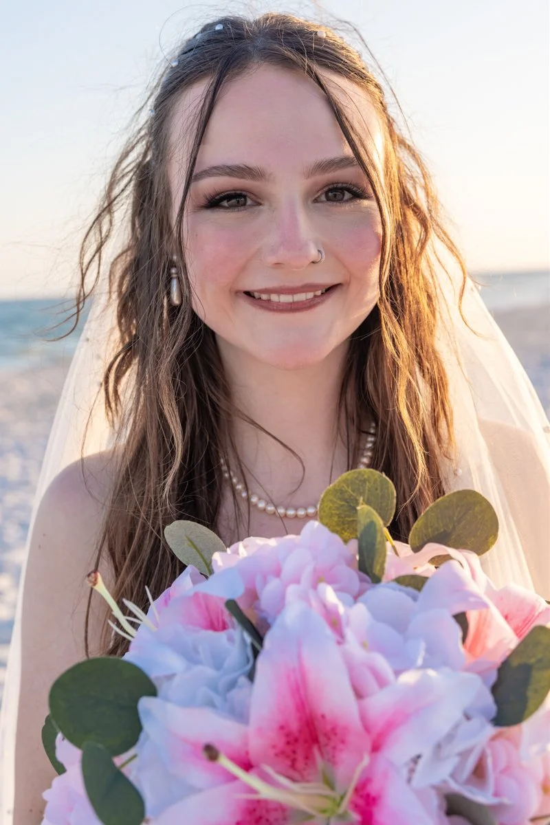 A young woman in a wedding dress holding a bouquet of pink and white lilies, smiling on a beach during sunset.