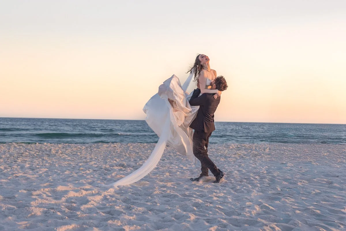 A man in a suit lifting a woman in a wedding dress on a beach at sunset, both laughing and enjoying the moment.