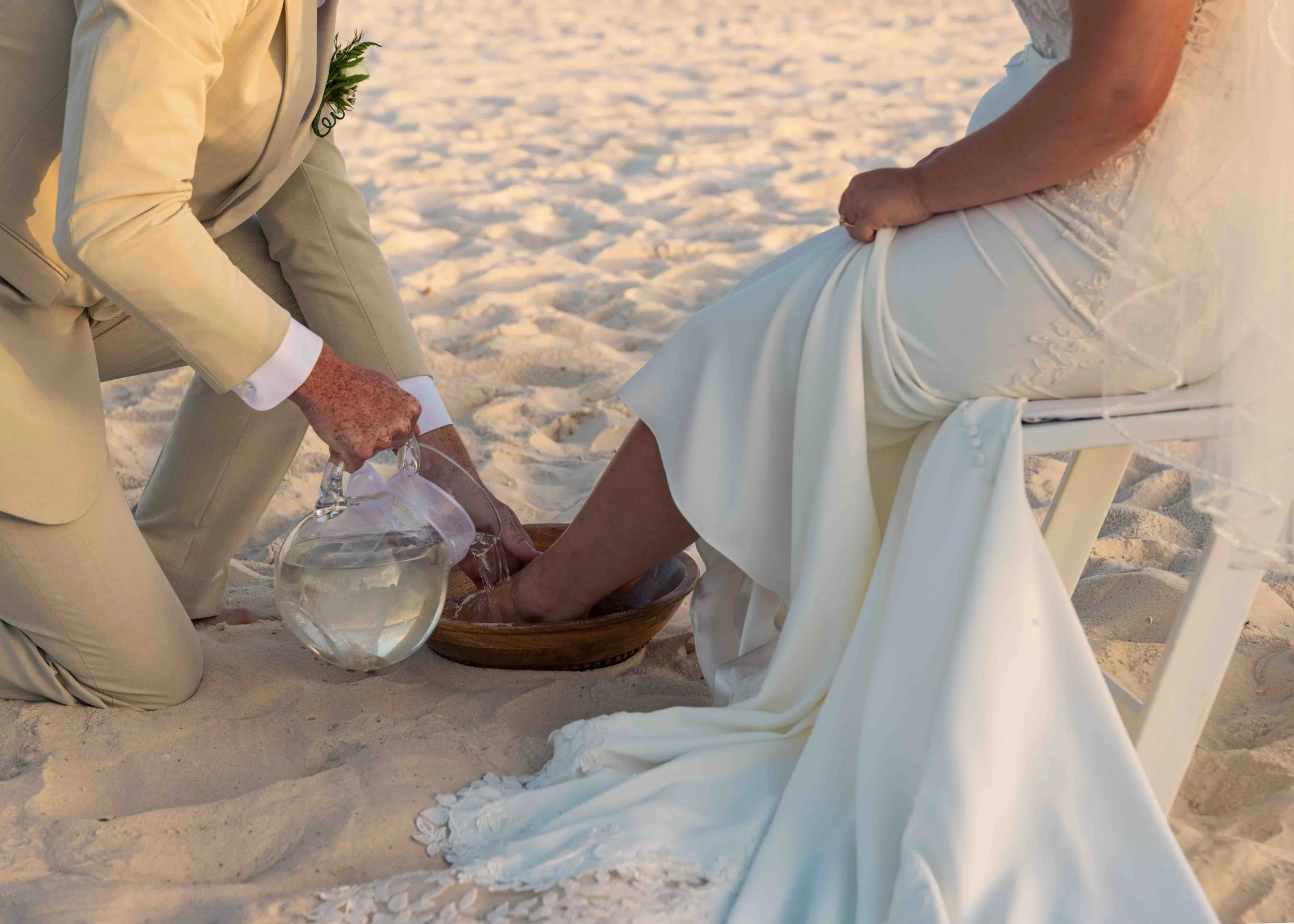A wedding ceremony on the beach where a man in a beige suit is kneeling and tying the shoe of a woman in a white wedding dress. They are on sand with minimal background visible.