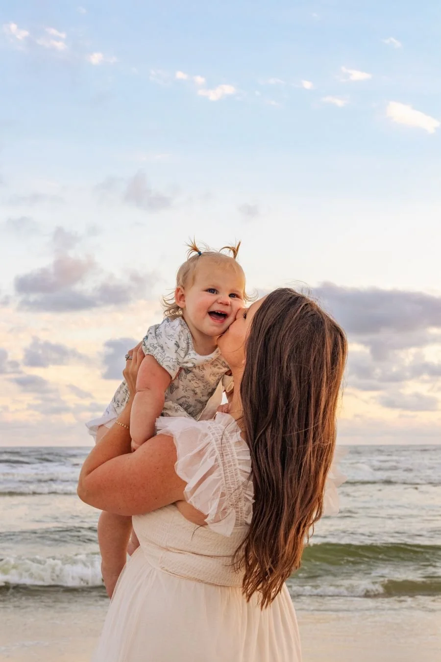 A woman holding a crying child at the beach during sunset, with ocean waves and a partly cloudy sky in the background.