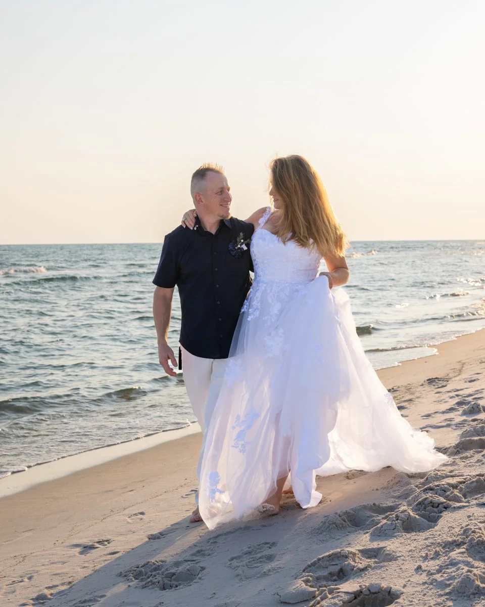 A bride and groom walking along the beach at sunset, the bride wearing a white wedding dress and the groom in a black shirt and white pants, ocean waves in the background.