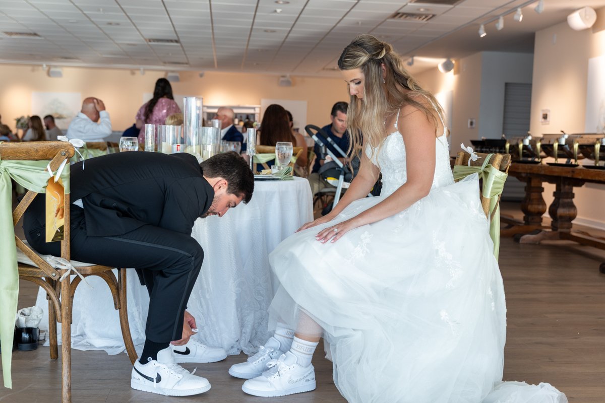 A bride in a white wedding gown sitting on a chair while a man in a black suit kneels to tie his shoelaces, both wearing white sneakers, at a wedding reception.