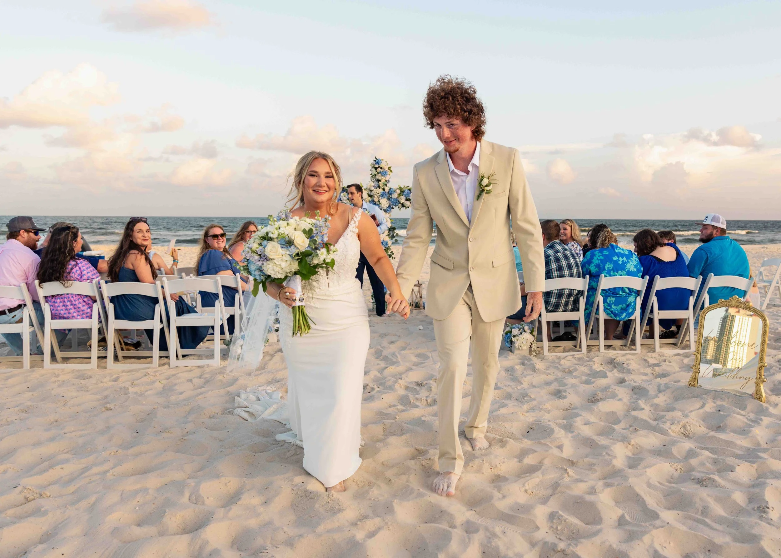 A bride and groom holding hands and walking on the beach during their wedding ceremony, smiling with guests seated in white chairs behind them, ocean and sky in the background.