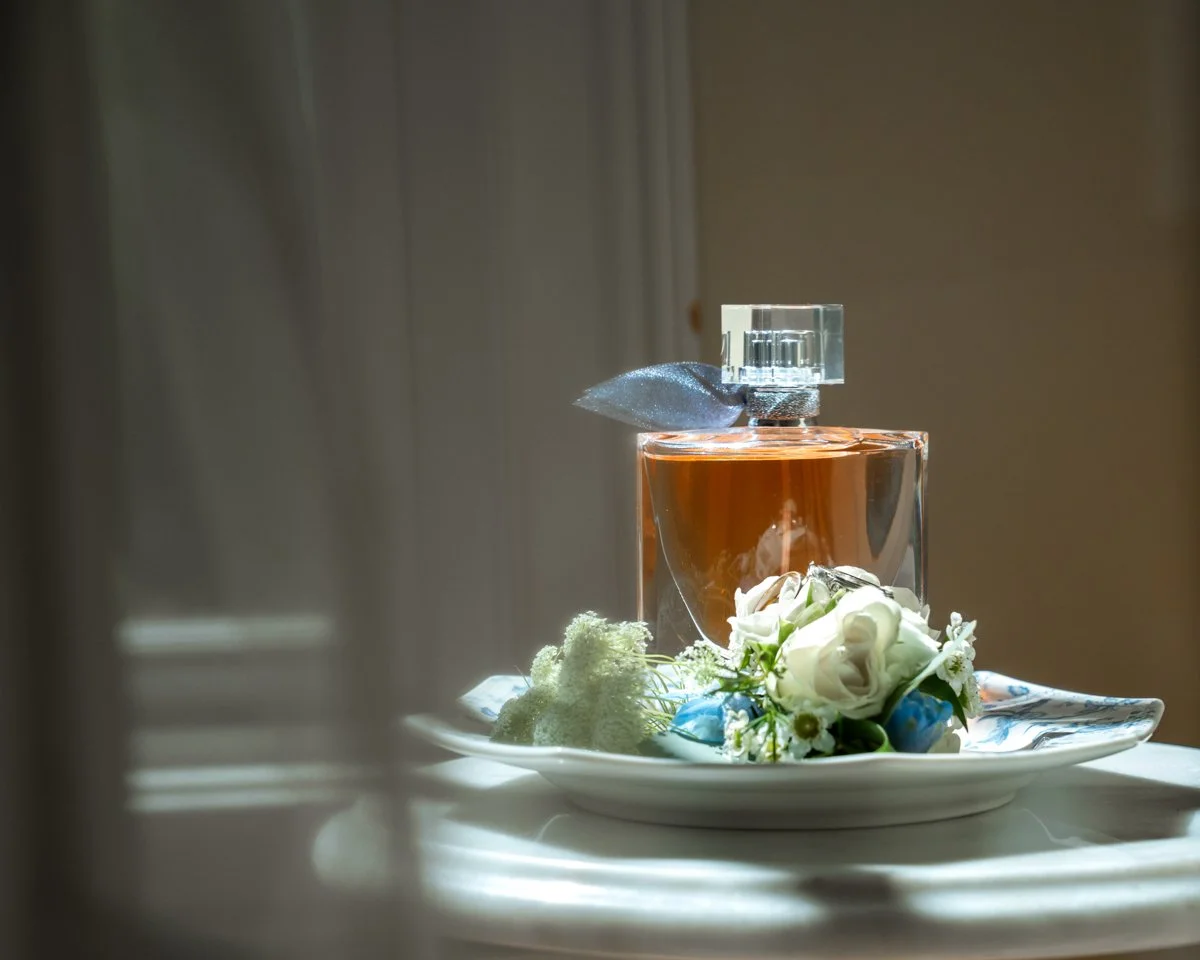 A glass perfume bottle with a transparent rectangular cap, adorned with a fabric ribbon, placed on a decorative plate with a floral arrangement of white roses and greenery.