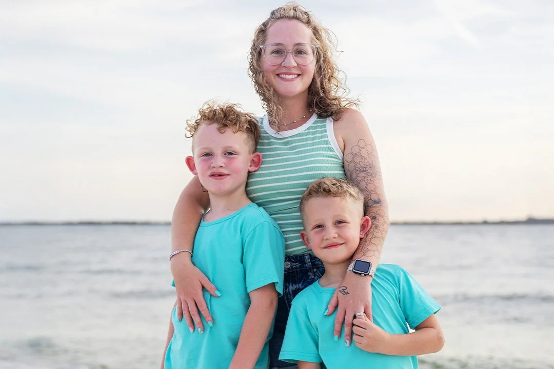 A woman with curly hair wearing glasses and a striped tank top stands on the beach with two young boys, all smiling and facing the camera.