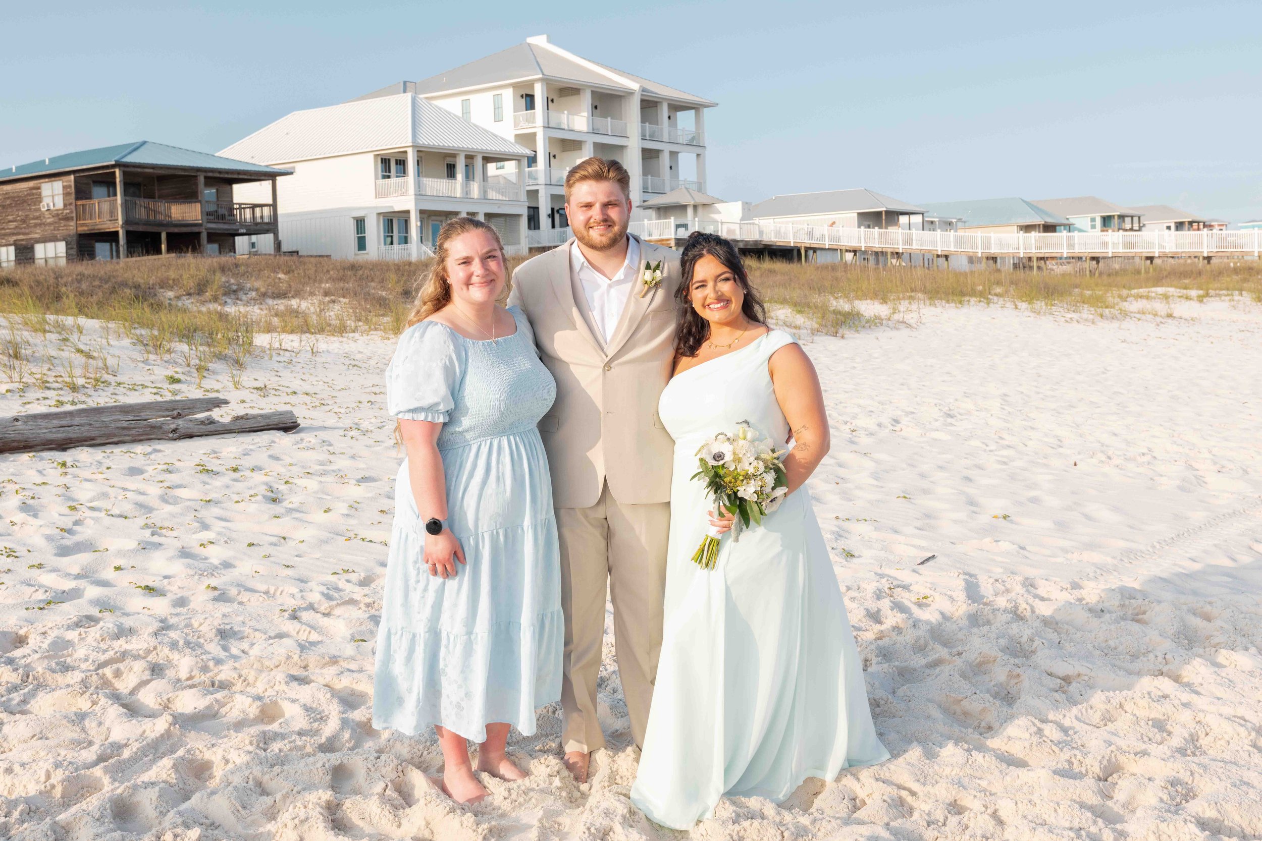 A group of three people, two women and one man, standing on a sandy beach with houses in the background, dressed in wedding attire, smiling, with one woman holding a bouquet of flowers.