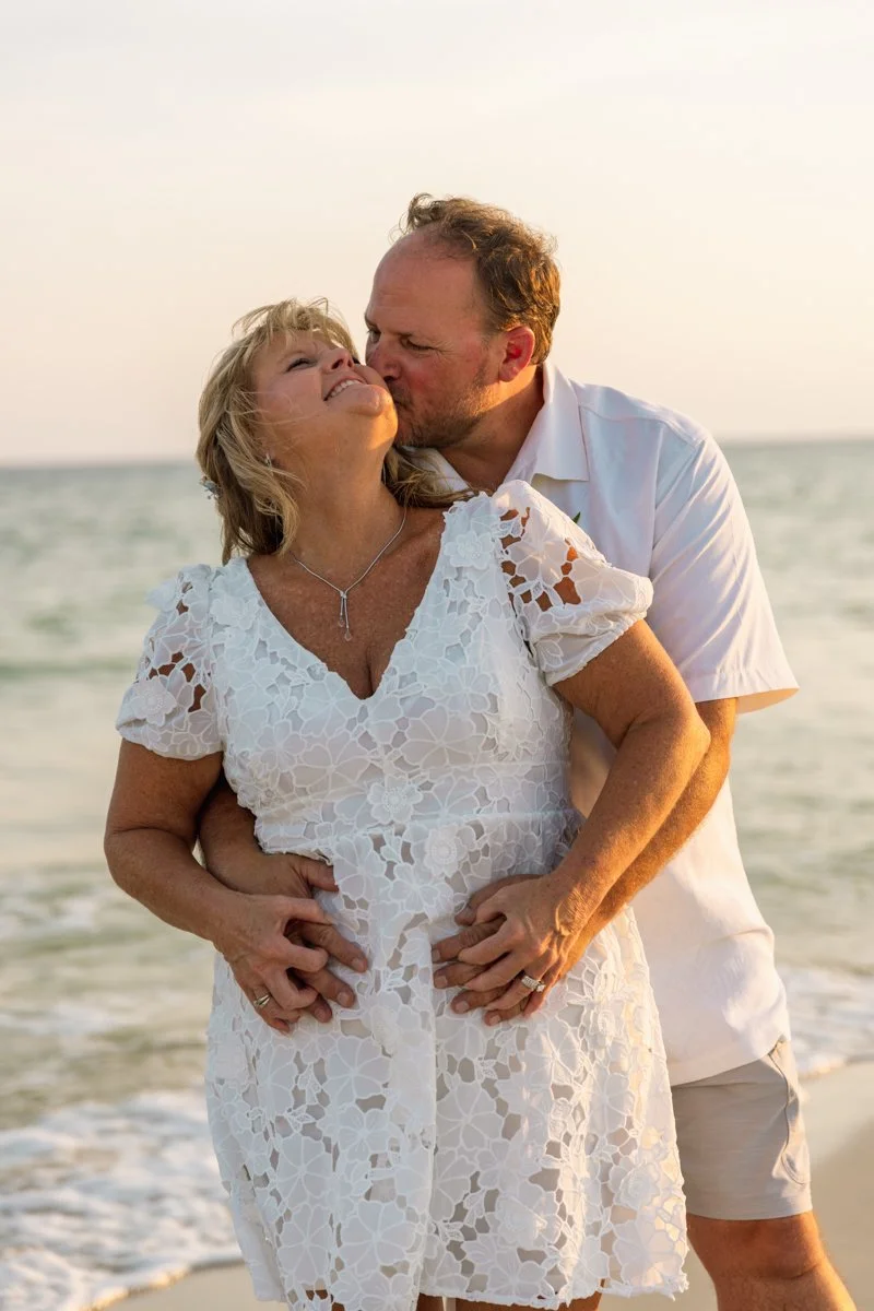 A man and woman embracing on the beach, with the man kissing the woman's cheek during sunset.