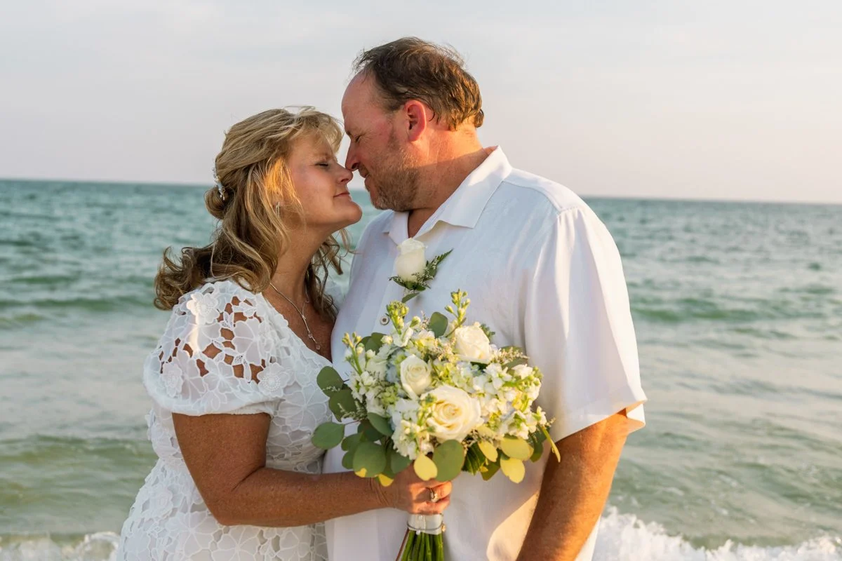 A couple in wedding attire embraces on a beach, with the ocean and sky in the background, holding a bouquet of white flowers.