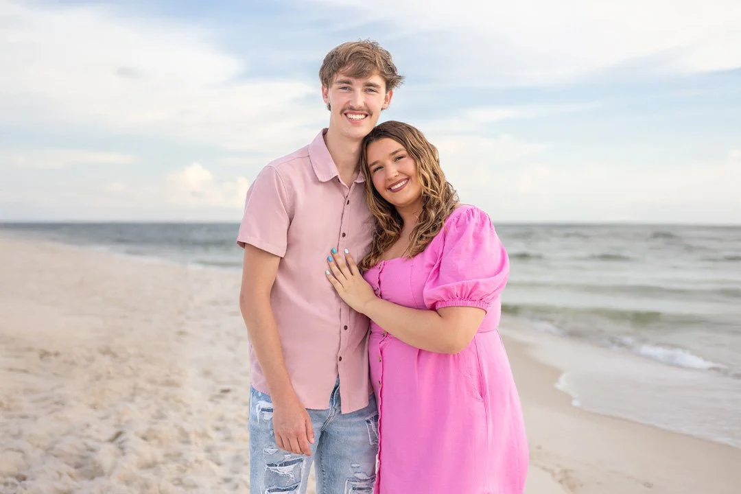 A smiling couple standing on a beach, embracing each other, with the ocean and sky in the background.