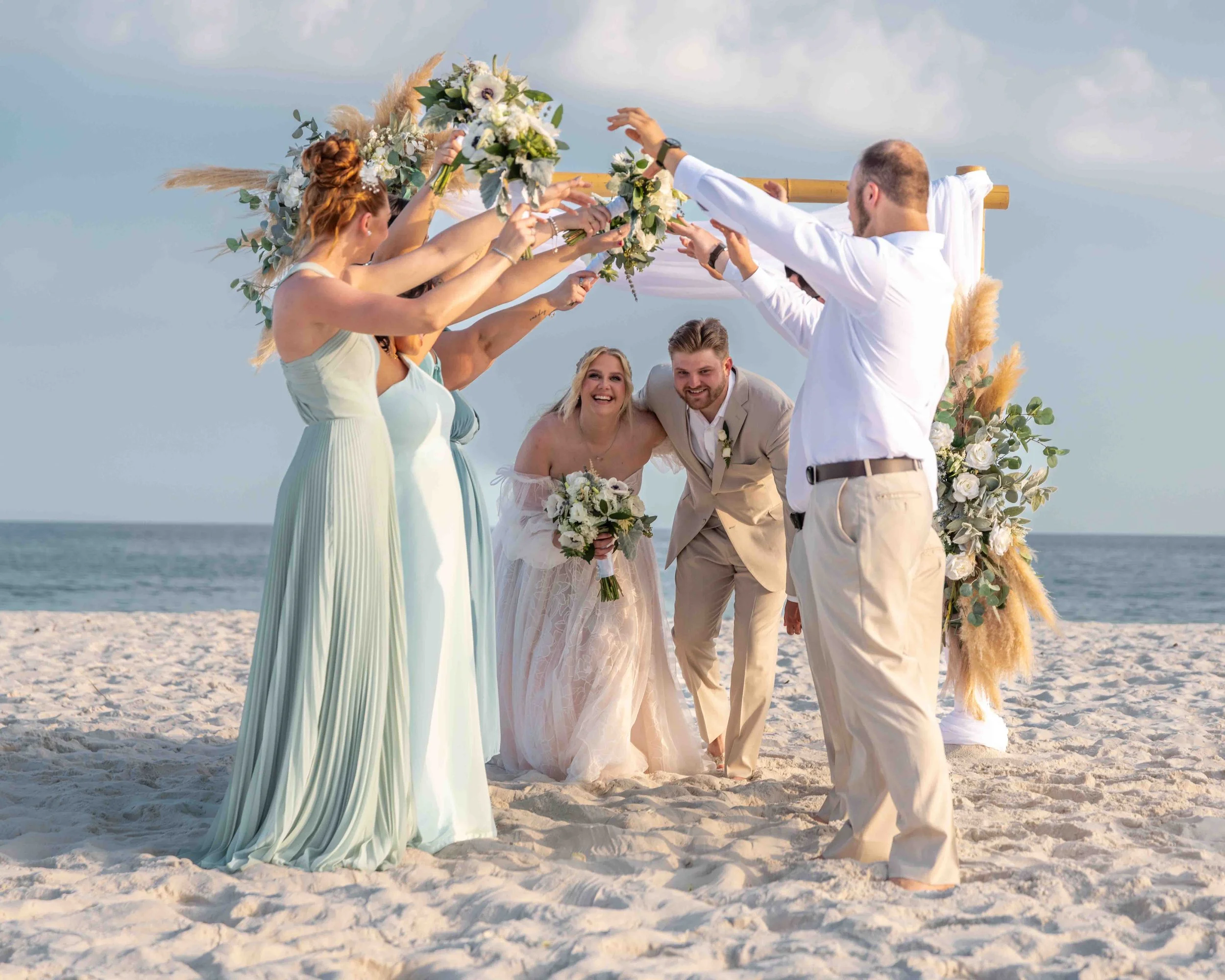 A wedding ceremony on the beach with the bride and groom surrounded by bridesmaids and groomsmen holding bouquets, with the ocean in the background.