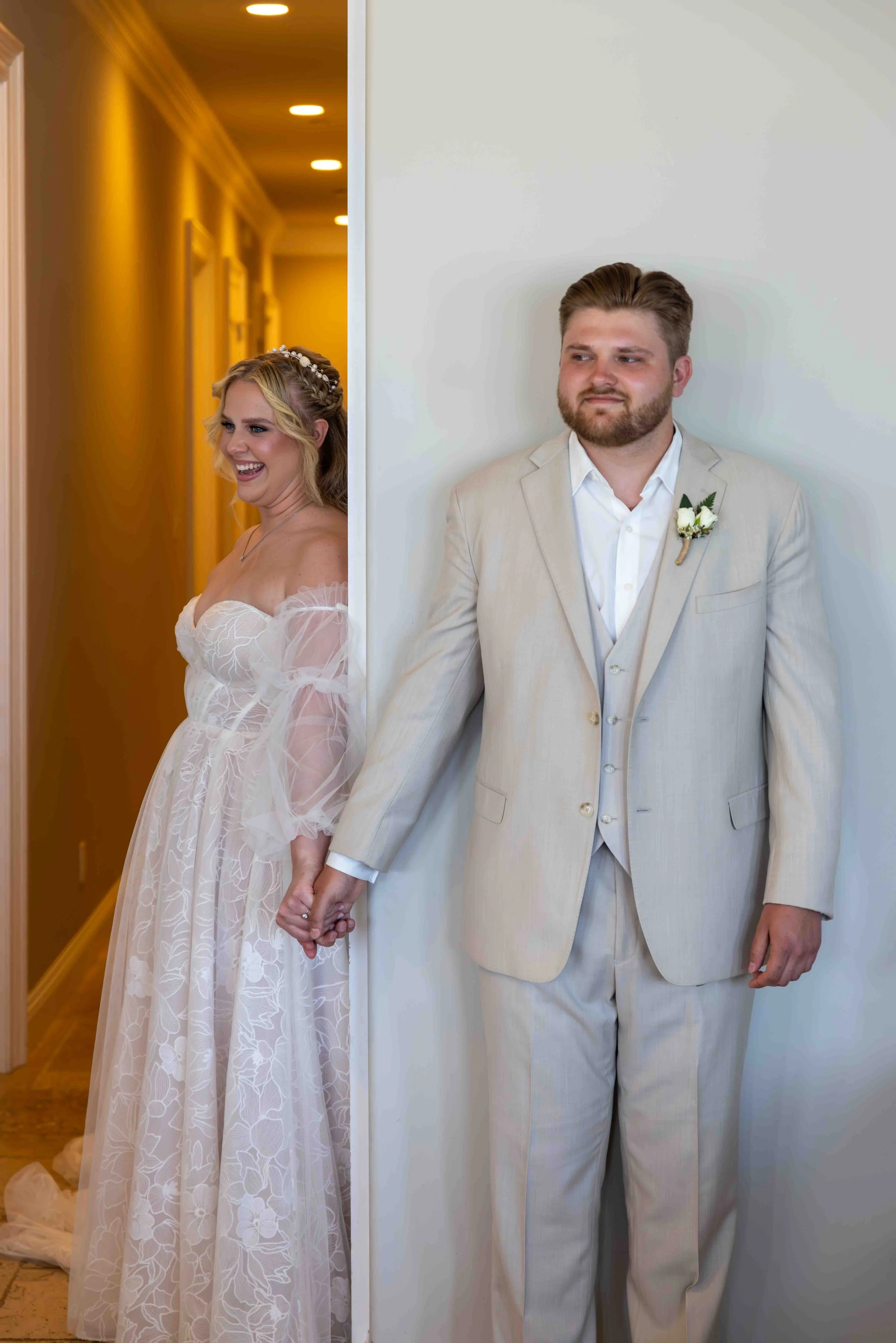 A bride and groom holding hands stand on opposite sides of a wall, with the bride smiling and the groom looking serious.