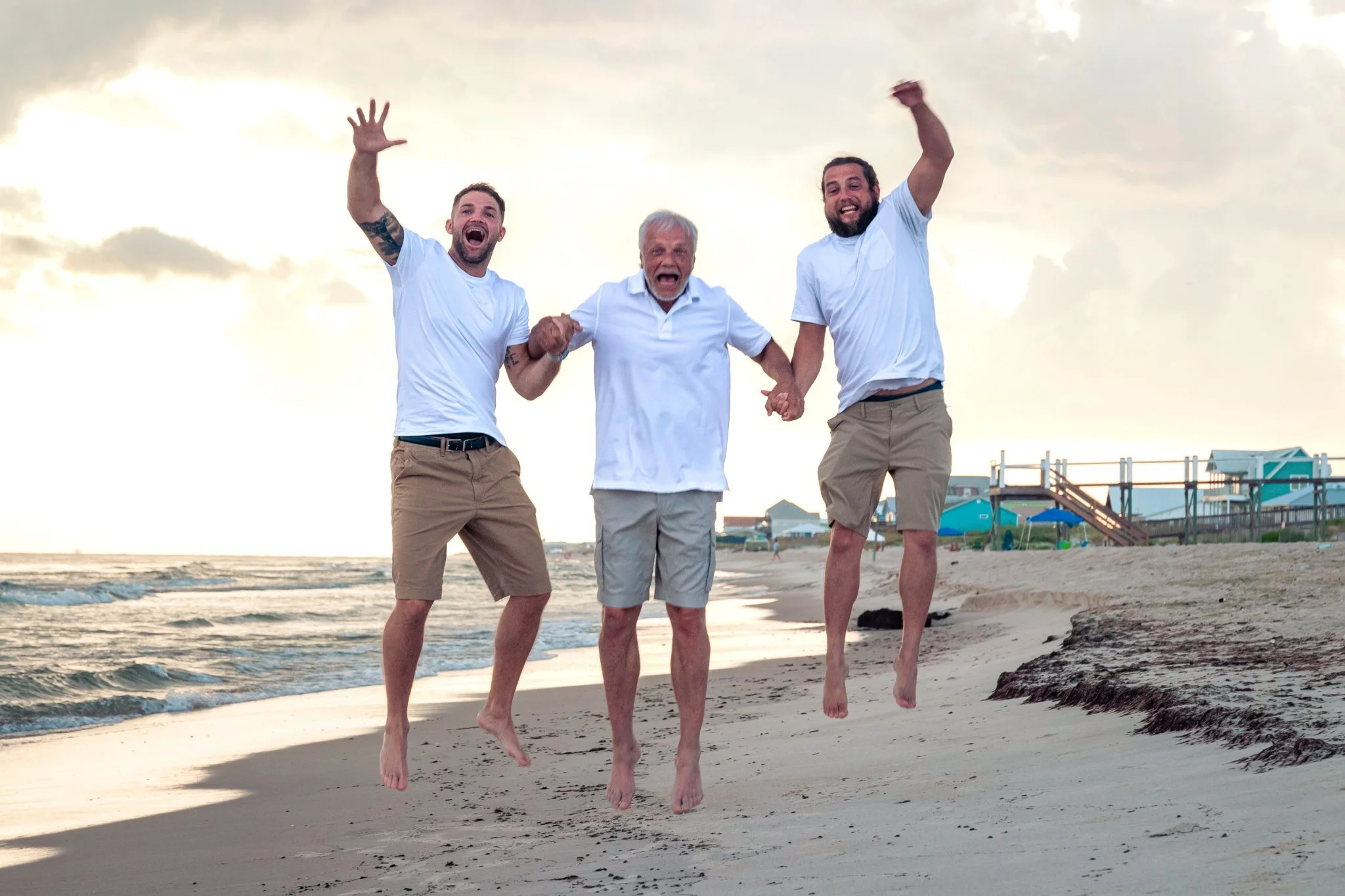 Three men jumping on the beach, holding hands, with the sky and ocean in the background. The man in the middle is older with gray hair, wearing a white polo shirt. The two men on either side are younger, wearing white t-shirts and beige shorts.