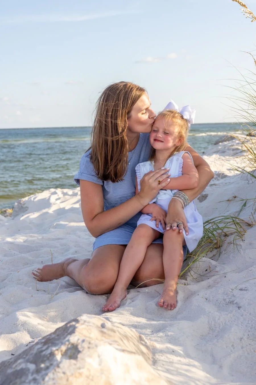 A woman and a young girl sitting on the sandy beach with the ocean in the background. The woman is giving the girl a kiss on the head, and the girl appears to be crying. The woman wears a blue shirt and the girl wears a white dress with a large white bow in her hair.