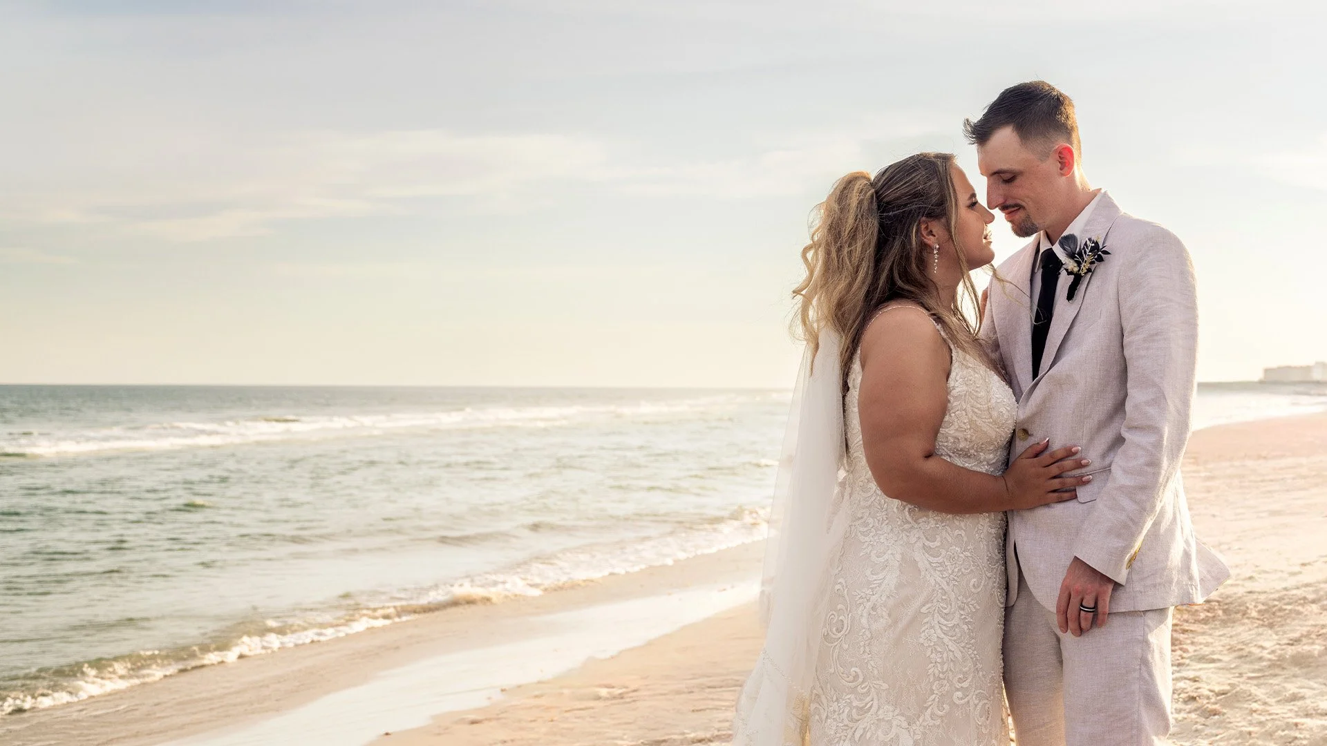 A bride and groom in wedding attire standing on a beach at sunset, holding each other and smiling with the ocean and sky in the background.