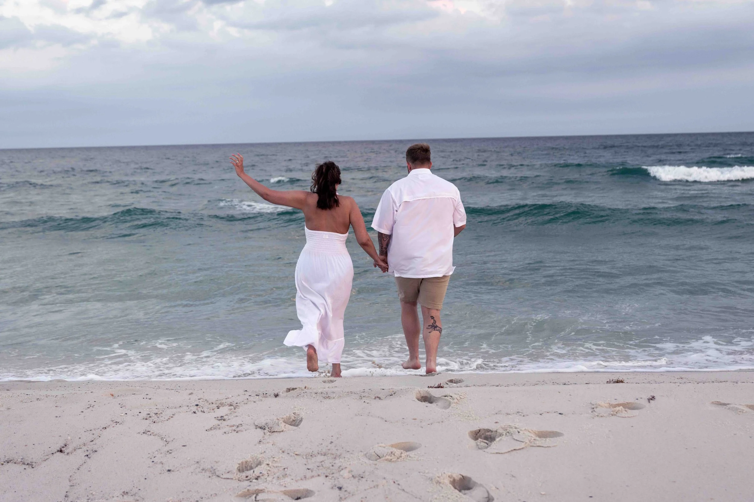 A couple walking hand in hand along the beach, with waves in the ocean and a cloudy sky above.