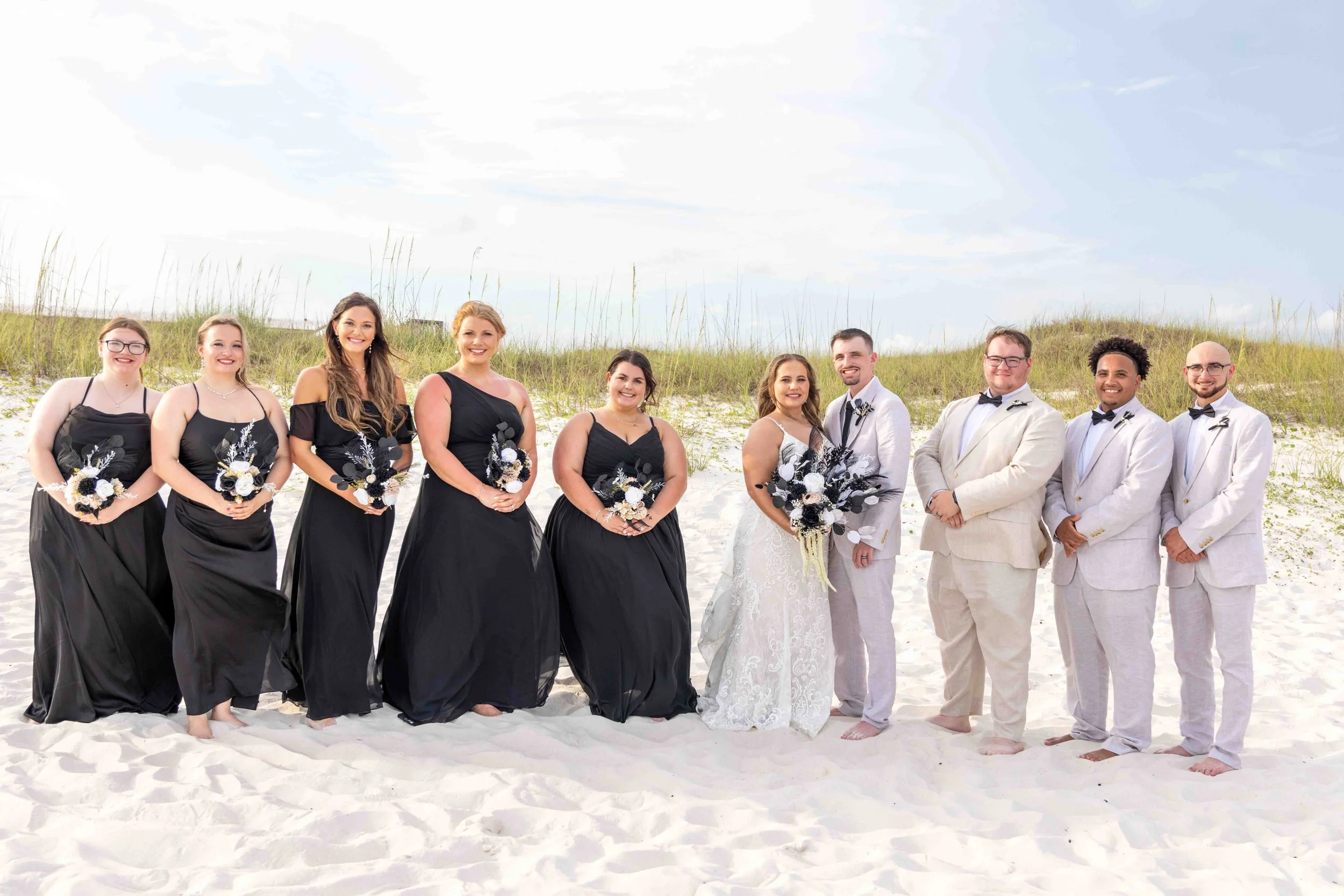A wedding party standing on a sandy beach with grass and blue sky in the background. The group includes women in black dresses and men in off-white suits with bow ties, all smiling at the camera.