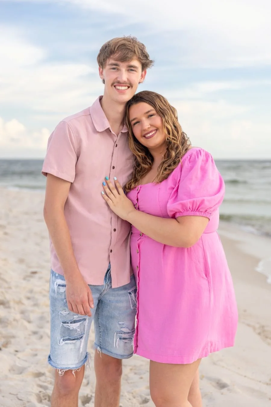 A young couple stands arm-in-arm on a beach, smiling; the man wears a pink shirt and ripped denim shorts, the woman in a pink dress.
