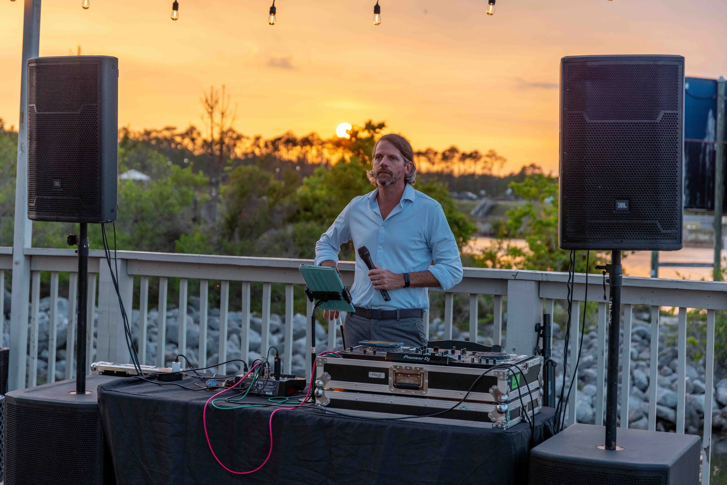 A man with long hair and a beard standing outdoors at sunset, holding a microphone. He is behind a DJ booth with audio equipment, speakers, and wires. There are string lights above and trees in the background.