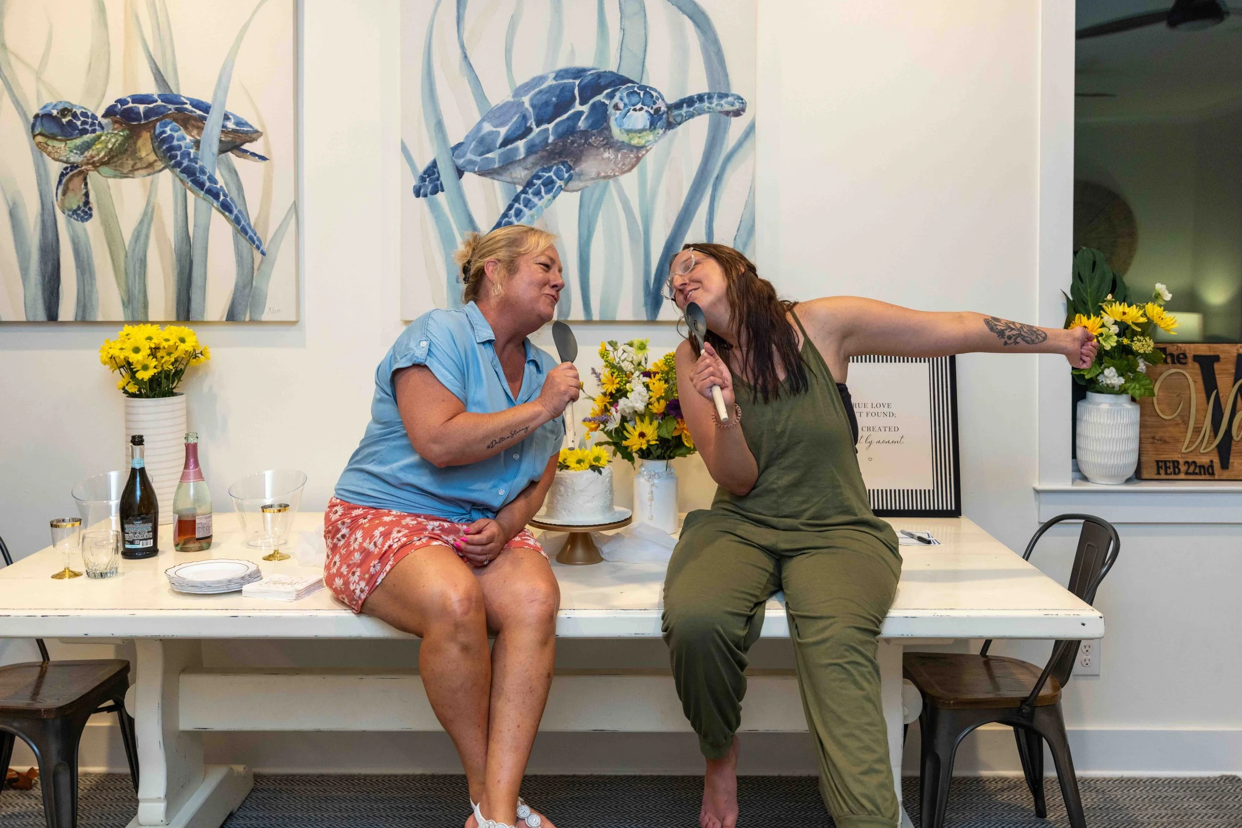 Two women are seated at a table, sharing a joyful moment and holding utensils. The woman on the left has blonde hair, is wearing a light blue shirt and floral shorts, and is smiling at the woman on the right. The woman on the right has brown hair, gl