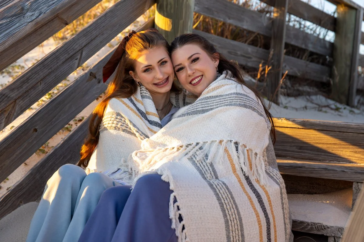 Two young women sitting together on a beach, wrapped in a white and gray striped blanket, smiling at the camera during sunset.