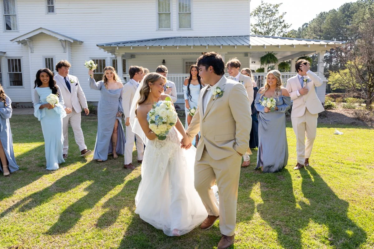 A wedding celebration outdoors with a bride and groom dancing in front of friends and family on a grassy lawn in front of a white house.