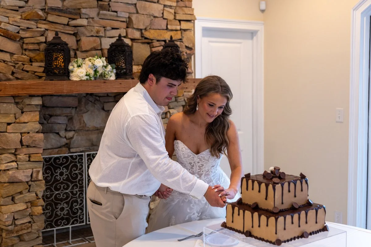 A bride and groom in wedding attire cutting a two-tiered wedding cake decorated with chocolate and placed on a white table.
