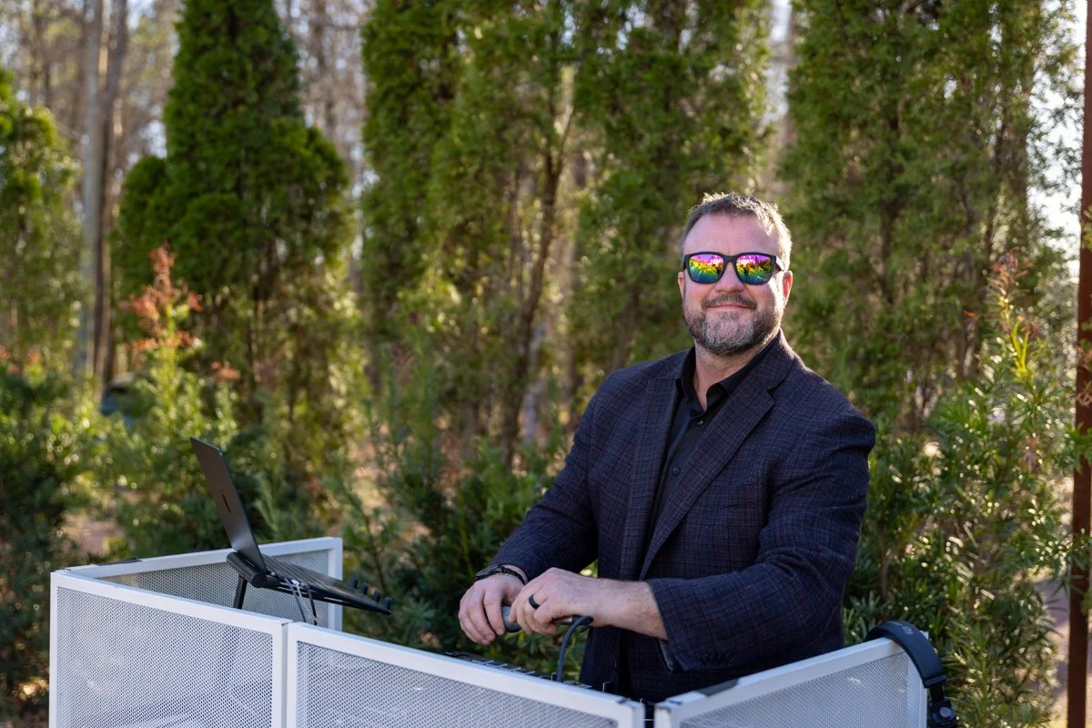 A man with a beard and sunglasses standing outdoors behind a DJ booth with a laptop, surrounded by greenery and trees.