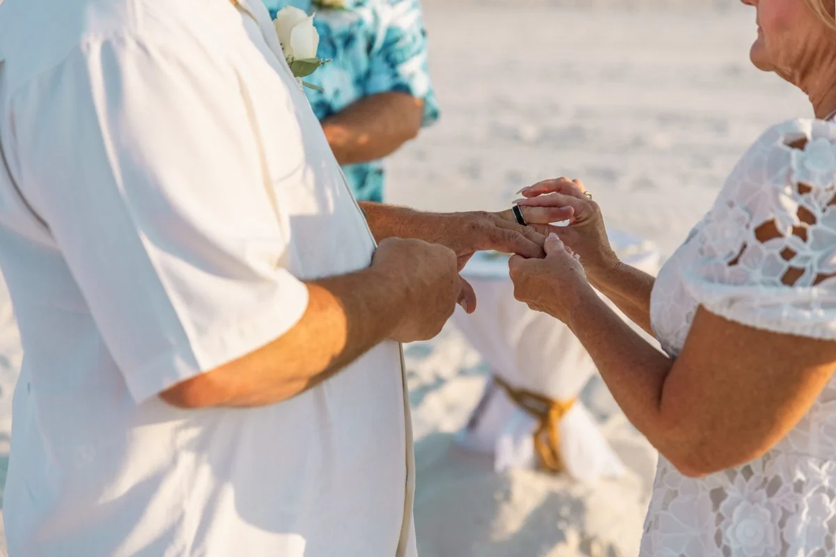 A couple exchanges rings during a beach wedding ceremony, with an officiant in the background.