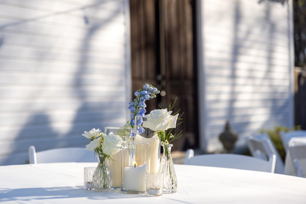 Table decorated with white roses, blue flowers, candles, and small glass votives outside in front of a shed or barn.