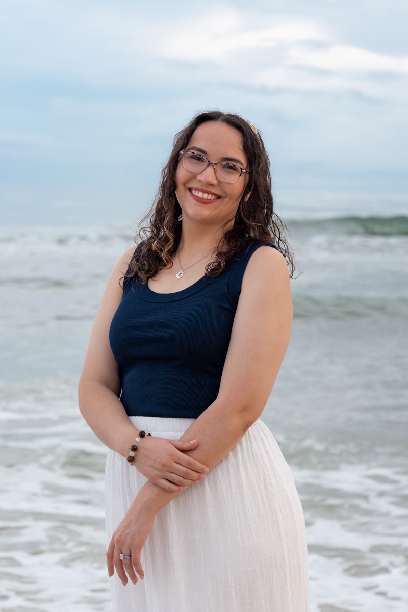 A smiling woman with glasses, wearing a navy blue sleeveless top and a long white skirt, standing on the beach with ocean waves and cloudy sky in the background.