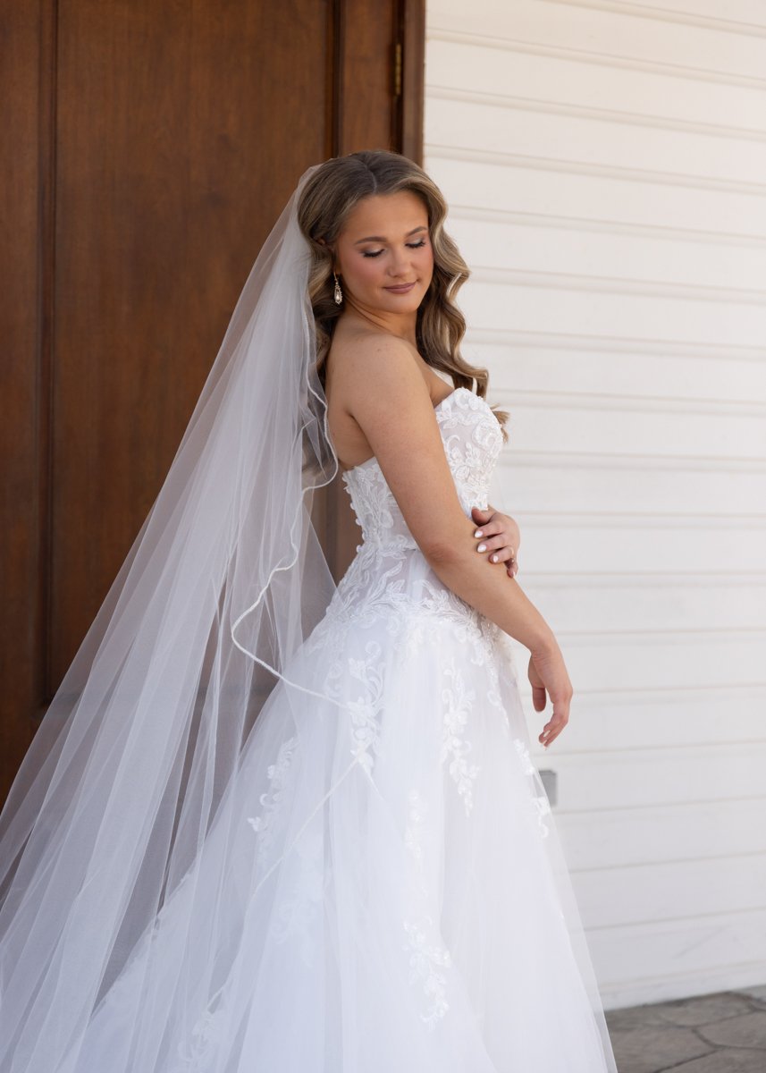 A bride in a white wedding gown and veil stands indoors, looking down with a gentle expression, in front of a wooden door and white panel wall.