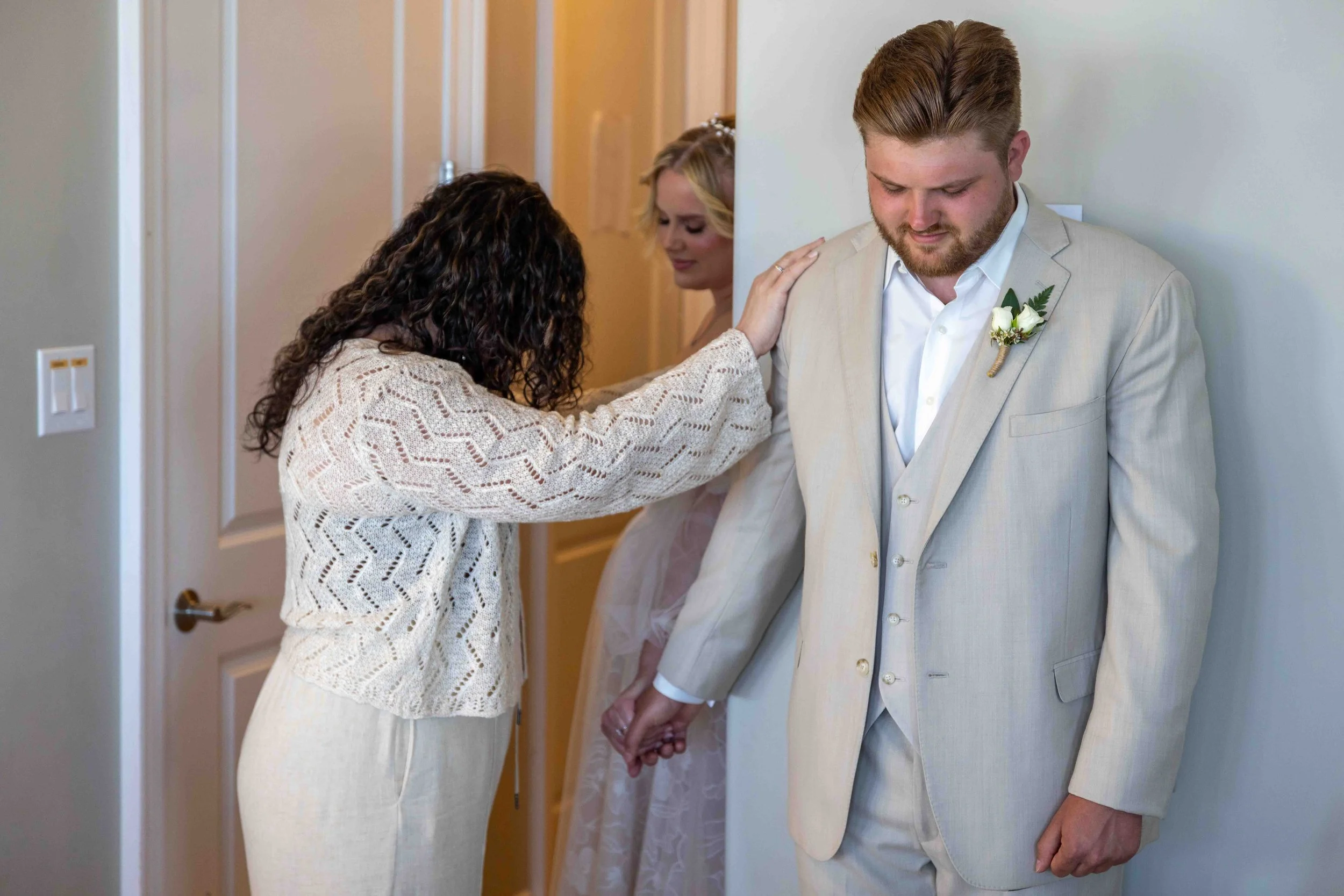 A couple holding hands, with the woman touching the man's shoulder, standing against a wall. The man wears a beige suit with a boutonniere, and the woman wears a cream dress.