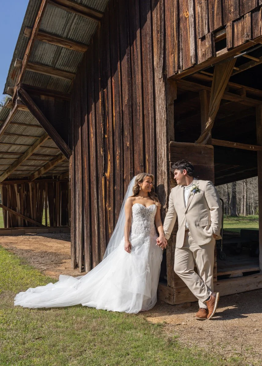 A bride and groom holding hands, standing outside a rustic wooden barn. The bride is wearing a strapless white wedding gown with a long train and a veil. The groom is dressed in a beige suit with brown shoes. They are smiling at each other in a sunny