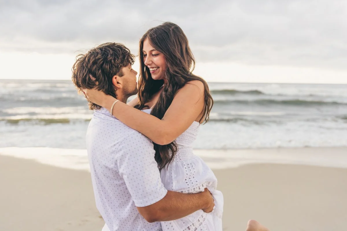 A couple on the beach during sunset, with the woman smiling and holding the man, who is looking at her.